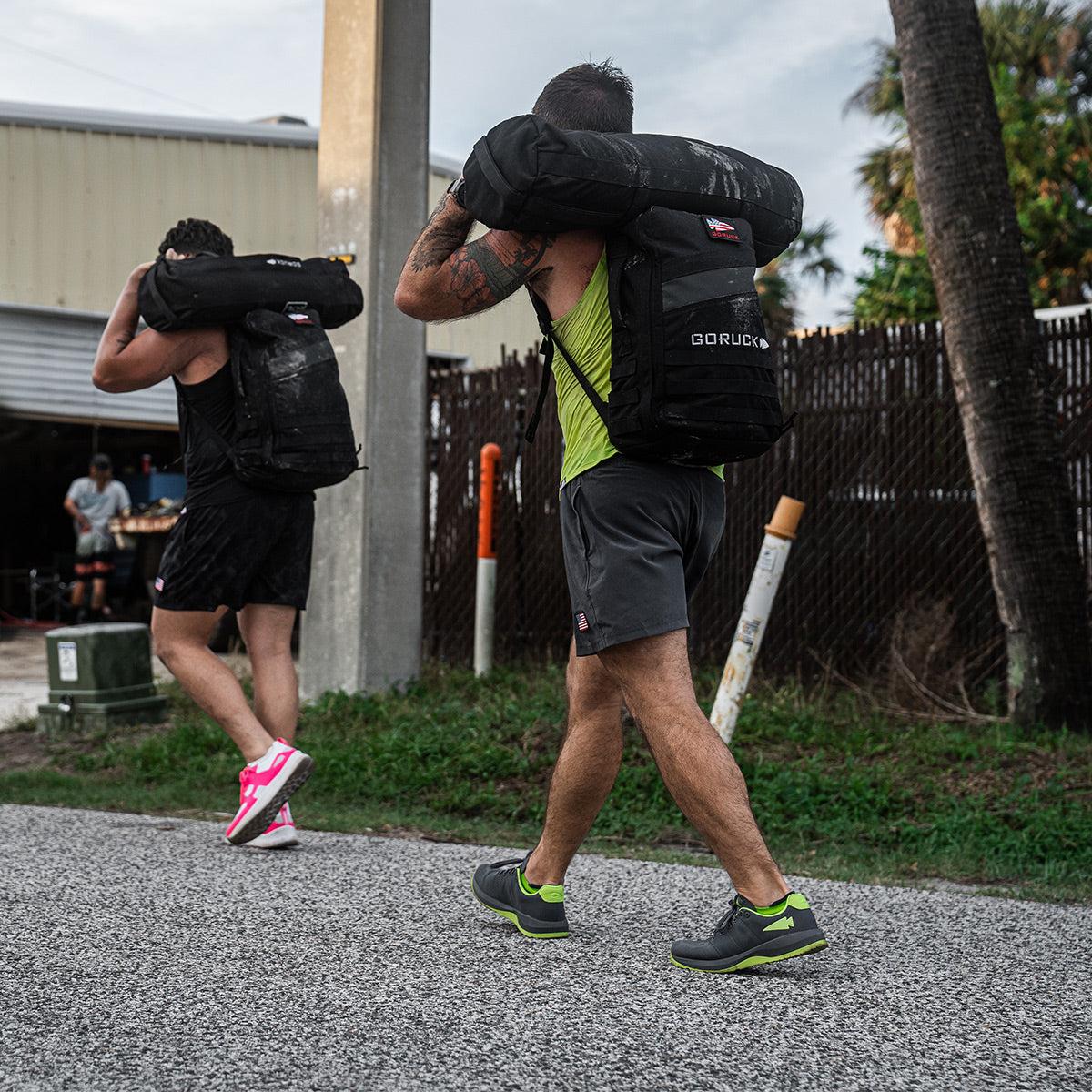 Two individuals outfitted in GORUCK's Men's Ballistic Trainers in Wolf Grey with Acid Lime accents and functional fitness attire carry large weighted sandbags crafted from CORDURA Ballistic Nylon on their shoulders as they walk outdoors. The scene unfolds on a gravel path near a fenced area adorned with palm trees and a partially visible building.