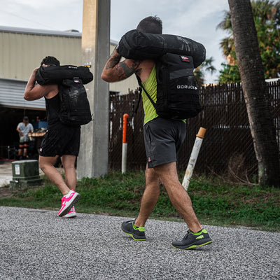 Two individuals outfitted in GORUCK's Men's Ballistic Trainers in Wolf Grey with Acid Lime accents and functional fitness attire carry large weighted sandbags crafted from CORDURA Ballistic Nylon on their shoulders as they walk outdoors. The scene unfolds on a gravel path near a fenced area adorned with palm trees and a partially visible building.