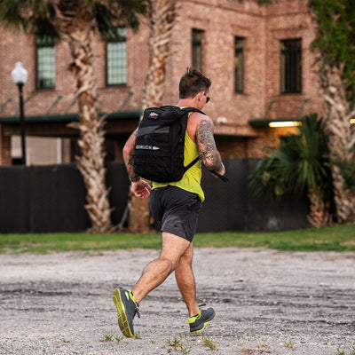 A person clad in a neon yellow tank top and black shorts strides along the gravel pathway in GORUCK's Men's Ballistic Trainers, featuring a Wolf Grey and Acid Lime design with an Acid Lime Reflective Spearhead. An accompanying durable backpack crafted from CORDURA Ballistic Nylon complements their run, while palm trees sway near a brick building in the background.