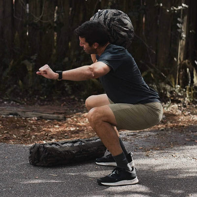 A person dressed in a dark shirt and khaki shorts confidently executes a squat, shouldering a durable camouflage bag that exemplifies the reliability assured by Scars Lifetime Guarantee. They are outdoors on a paved area amidst trees and foliage, with another bag resting on the ground nearby.