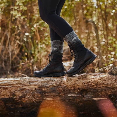 A person wearing black hiking boots and cozy GORUCK Merino Challenge Socks 1.0 - Crew walks on a fallen log in a forest. Sunlight filters through the trees in the background, casting a warm glow.