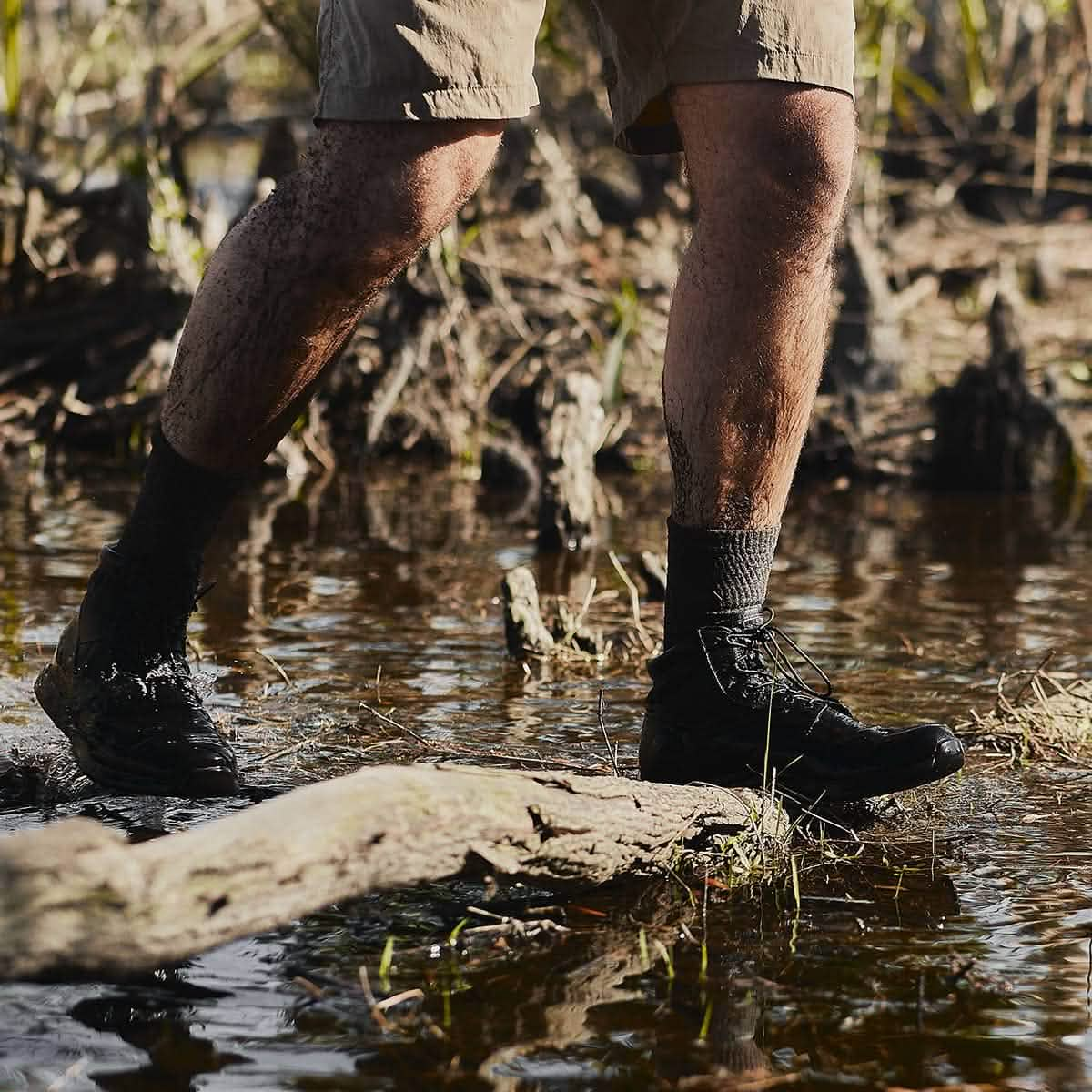 A person wearing shorts, hiking boots, and GORUCK's Merino Challenge Socks 1.0 - Crew steps onto a muddy log in a swampy area, surrounded by water and vegetation. The boots and legs are splattered with mud, signaling a rugged outdoor adventure backed by Scars Lifetime Guarantee.