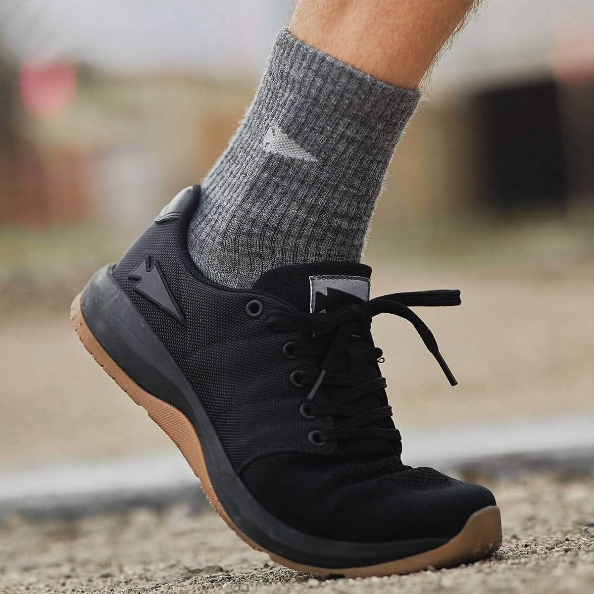 A close-up image of a person's foot wearing a black sneaker with brown soles and GORUCK's Merino Challenge Socks 1.0 - Crew in gray. The shoe rests on a rough outdoor surface, while the merino wool socks, celebrated for their comfort and durability, display a small logo that signifies quality and style.