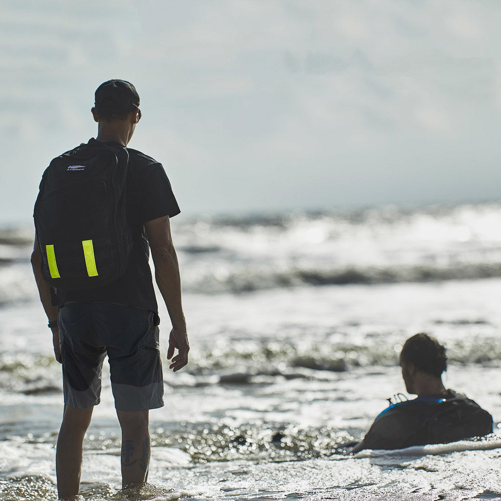 A person stands on the beach wearing a black t-shirt, Men’s Challenge Shorts by GORUCK with cargo pockets made from Lightweight ToughDry® fabric, and a backpack. Another sits in the water facing the ocean. The scene is calm with gentle waves and a cloudy sky.