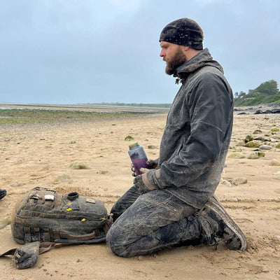 A person kneels on a sandy beach, holding a container. Dressed warmly in a beanie, gloves, and jacket with lightweight running shoes, they appear ready for any adventure. A GORUCK Men's Rough Runner - Black + White backpack lies beside them against the cloudy sky and distant shoreline, embodying the spirit of exploration.