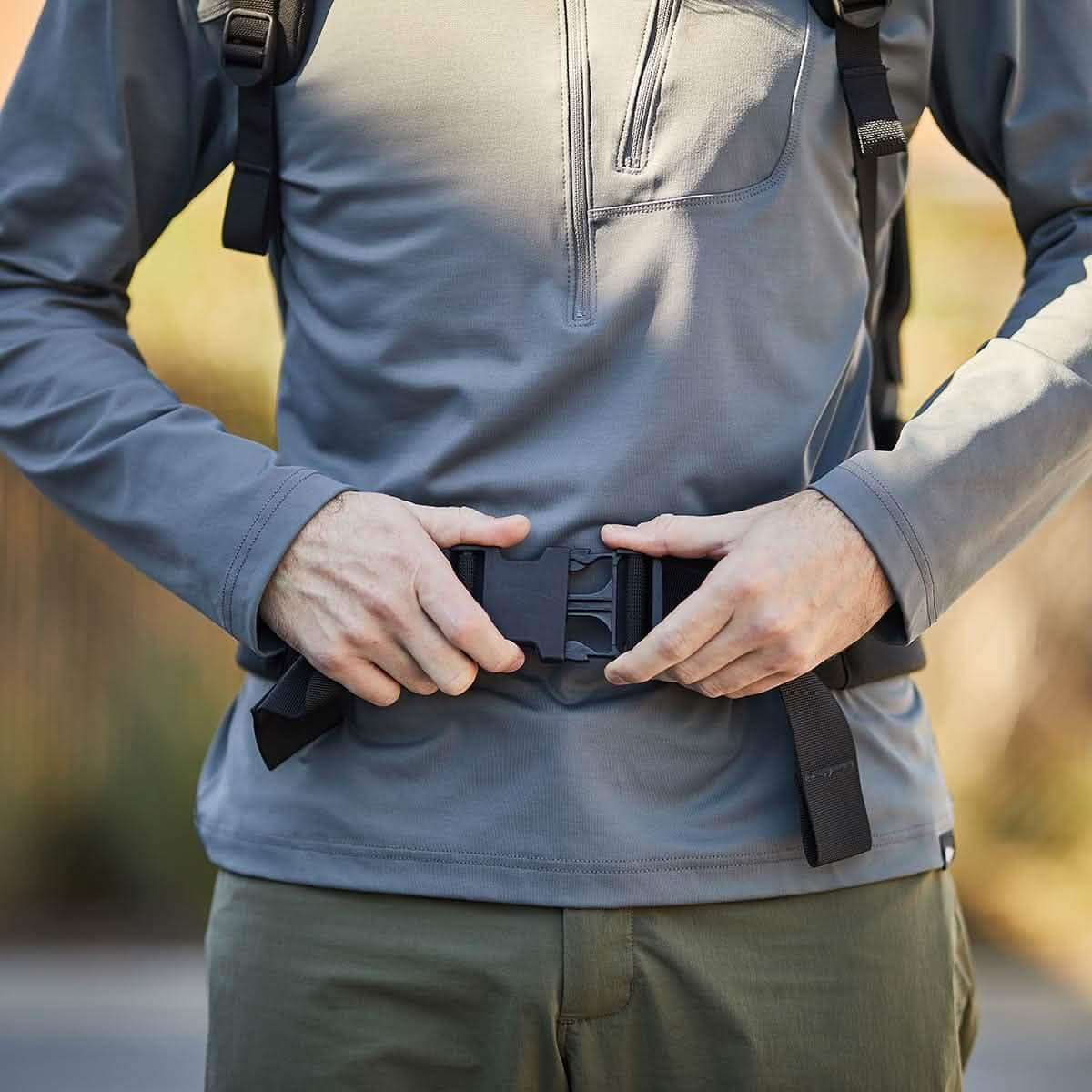 Outside, someone fastens a black belt buckle on a gray jacket with green pants, embodying rugged work ethos celebrated by the mikeroweWORKS Foundation and showcased in the GORUCK X mikeroweWORKS Bundle.