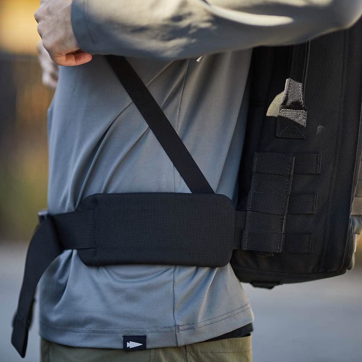 A person in a gray shirt adjusts the strap of the GORUCK X mikeroweWORKS Bundle backpack, featuring a black GR1 with a padded waist belt, perfect for rugged jobs.