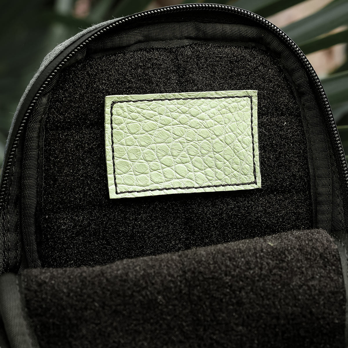 The interior pocket of a black backpack, adorned with an Everglades Mystery Leather Patch by GORUCK, showcases its exotic green texture. The open pocket is set against a softly blurred backdrop of green plant leaves, infusing the sophisticated design with a natural touch.