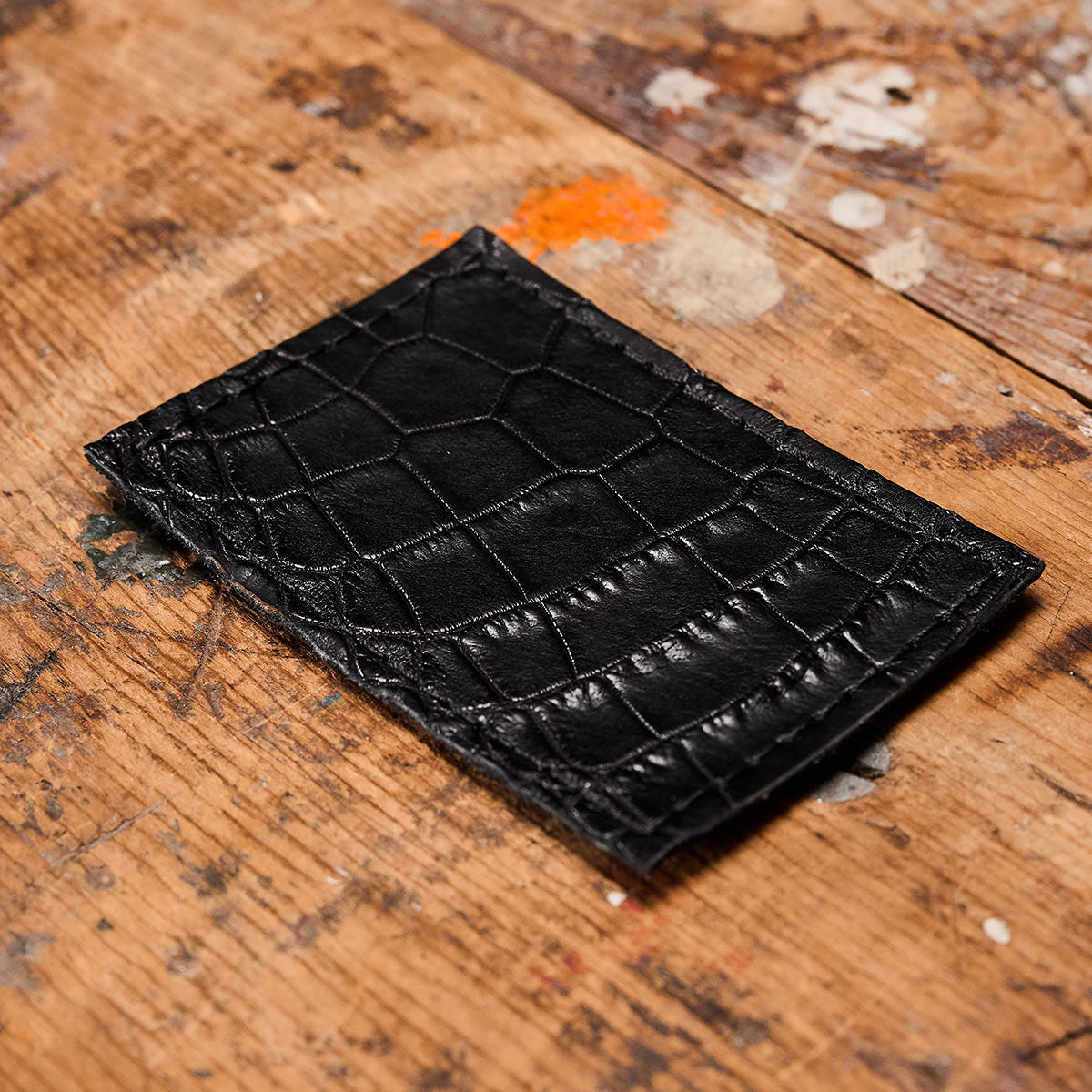 An Everglades Mystery Leather Patches wallet by GORUCK, designed with an exotic crocodile pattern, lies on a rustic wooden surface adorned with scattered paint stains in various colors, giving the scene an artistic flair.