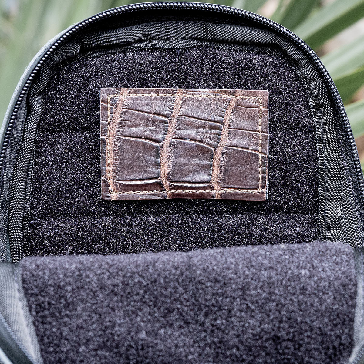 Close-up of an open backpack showcasing an Everglades Mystery Leather Patch by GORUCK, intricately crafted from exotic leather with a crocodile skin pattern on the inner lining. The background features a blurred view of green leaves.