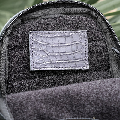 Close-up of an open black leather bag from GORUCK, featuring a textured crocodile leather patch sewn on the inner lining. Known as the Everglades Mystery Leather Patches - Built By Scars, this bag showcases its unique patches amid lush green leaves in the background.