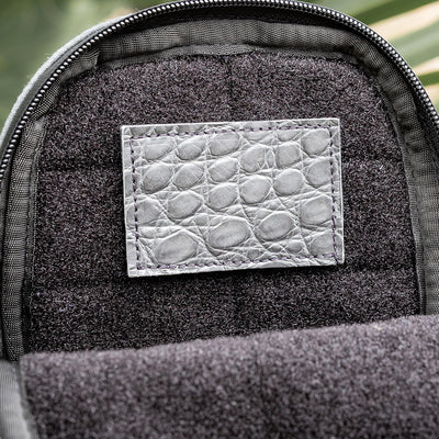 Close-up of a dark gray backpack interior featuring a unique Everglades Mystery Leather Patch by GORUCK, showcasing an exotic crocodile skin design. The leather patch is stitched onto the soft fabric lining, highlighted by natural lighting.