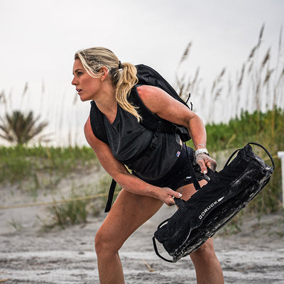 A woman in workout attire with a ponytail holds a sandbag and wears a backpack. She sports the Women’s USA Performance Tank - ToughMesh by GORUCK, which dries lightning fast. Positioned on a sandy beach with tall grass in the background, she appears focused and ready for exercise.