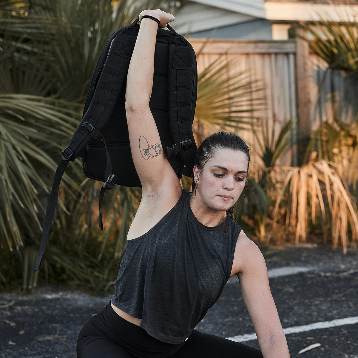 A woman in a Women’s USA Performance Tank - ToughMesh by GORUCK performs an outdoor exercise, lifting a black backpack above her head. Surrounded by palm trees and a wooden fence, she enjoys the comfort of the lightning-fast drying ToughMesh™ fabric.