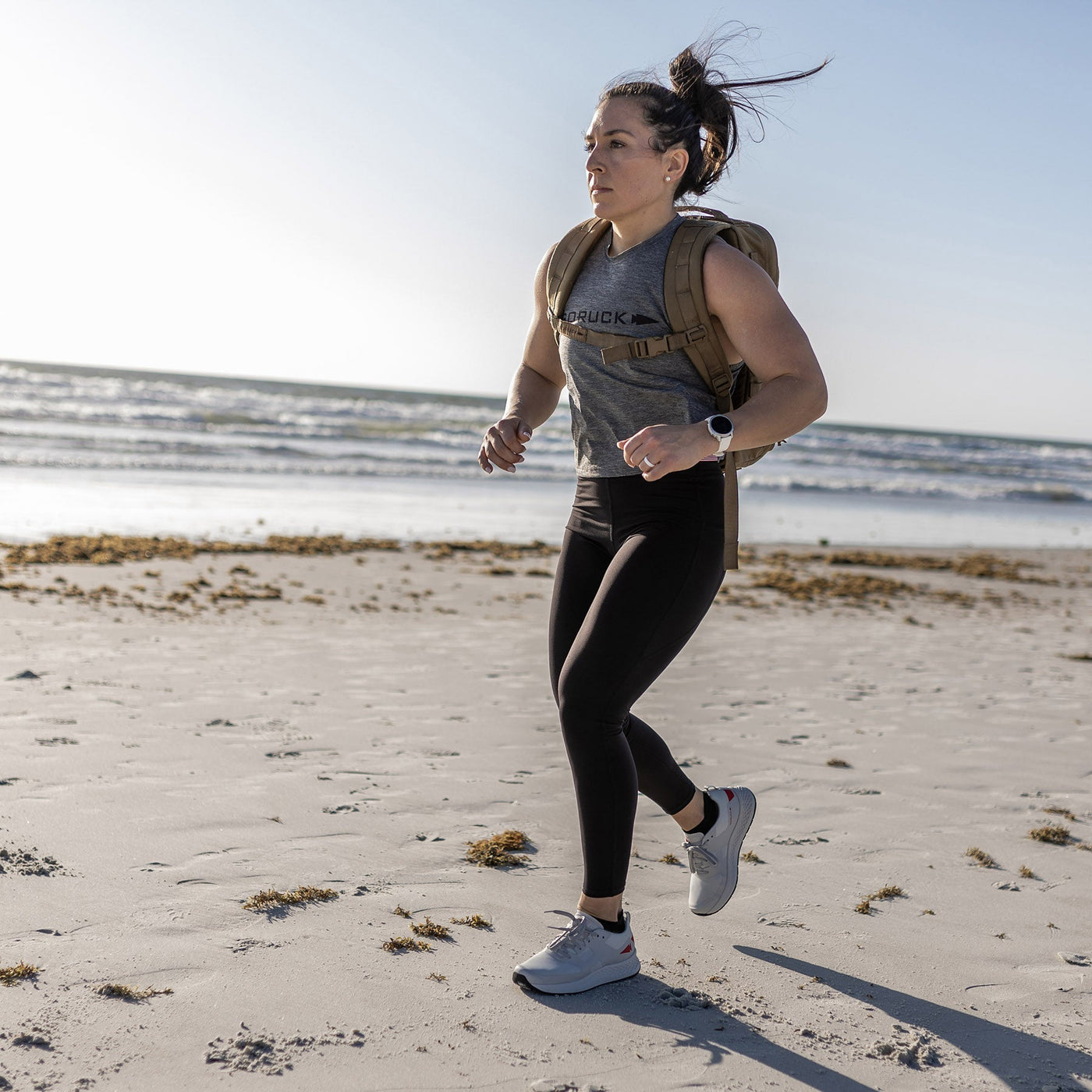 A woman jogs along a sandy beach with seaweed scattered on the shore. She's wearing high mileage Women's Rough Runner shoes in light grey and red by GORUCK, paired with a gray tank top and black leggings, while carrying a backpack. Her hair is tied back, and she checks her smartwatch as the ocean and blue sky stretch out in the background.