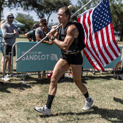 A person in athletic gear and Women's Rough Runner shoes from GORUCK carries a large American flag while running outdoors. Onlookers watch from behind a barrier displaying a banner that reads GORUCK Sandlot. The scene is sunny with trees in the background, capturing the spirit of a true Rough Runner.