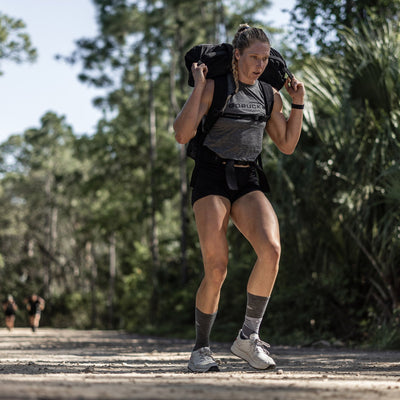 A person exercises outdoors, trekking along a dirt path surrounded by trees with a weighted backpack. They sport a gray tank top, black shorts, and the GORUCK Women's Rough Runner in Light Grey + Red, which features a Gradient Density EVA Midsole. Sunlight filters through the trees casting shadows on the path.