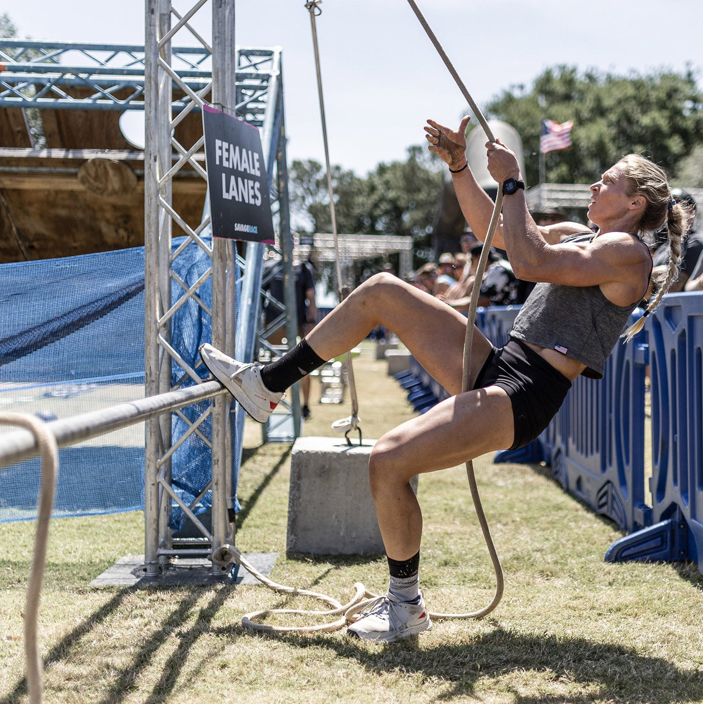 During the Rough Runner event, a participant equipped with GORUCK's Women's Rough Runner in Light Grey + Red ascends a rope next to a Female Lanes sign on the outdoor obstacle course. Her athletic gear captivates spectators watching from behind the barrier while an American flag gracefully waves in the background, symbolizing determination and spirit.