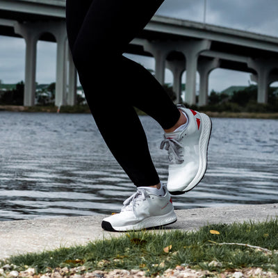 A person sporting the Women's Rough Runner - Light Grey + Red by GORUCK jogs along a riverbank on a cloudy day, with a large bridge looming in the background.