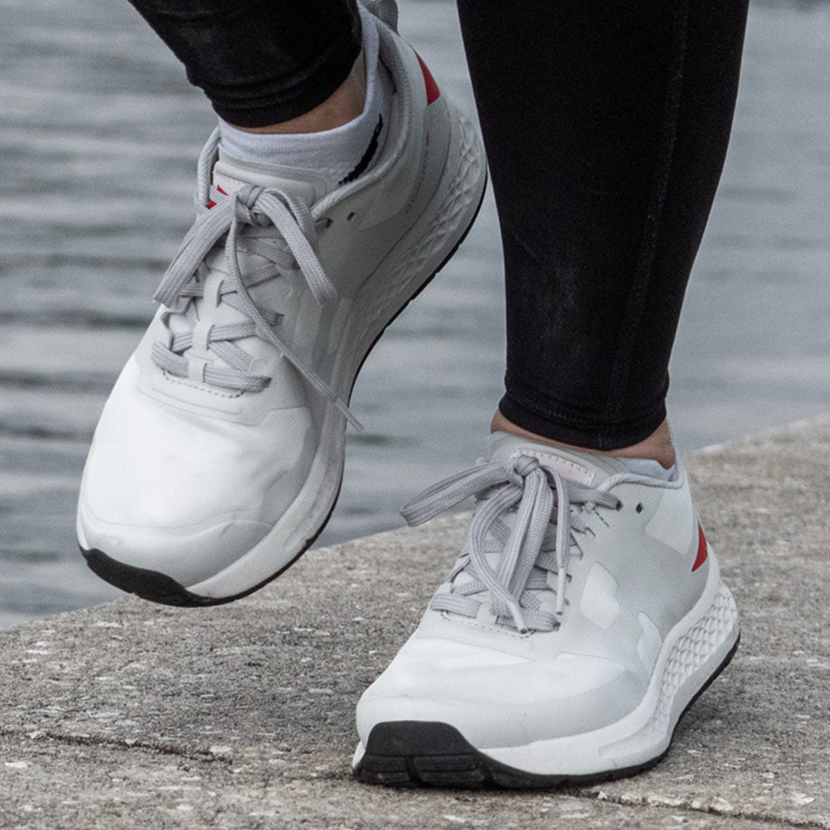 A close-up of a person wearing the Women's Rough Runner by GORUCK in Light Grey with Red accents, featuring a Gradient Density EVA Midsole, standing on concrete near water. They are paired with black leggings.