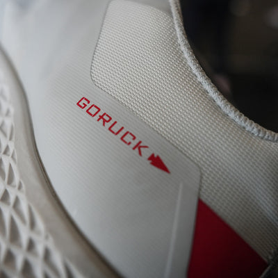 Close-up of a light grey sneaker showcasing the GORUCK brand name in red on the side. This shoe, known as the Women's Rough Runner - Light Grey + Red, features a textured sole and a Gradient Density EVA Midsole, perfect for high mileage running and outdoor activities.