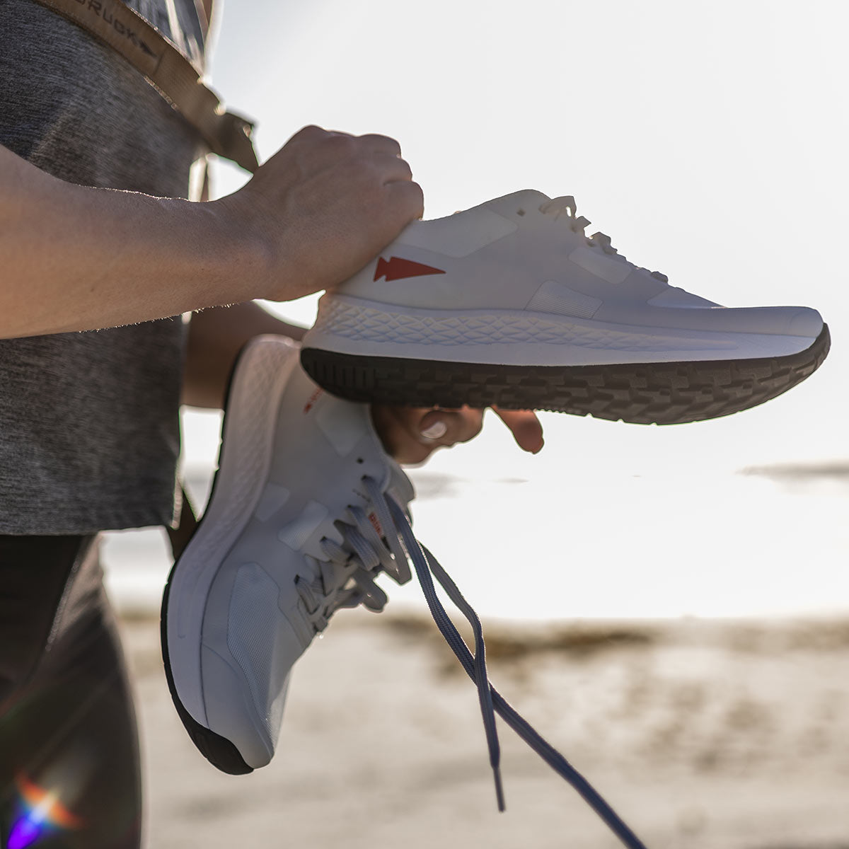 A person holding a pair of GORUCK Women's Rough Runner shoes in light grey with a red logo, standing on a beach with sunlight reflecting off the sand and water in the background.