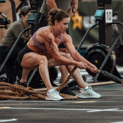 In a gym setting, a woman in athletic wear from the GORUCK Women's Rough Runner collection in Light Grey + Red showcases strength and determination as she pulls a thick rope. Others exercise amidst the equipment. Her focus mirrors that of preparing for a rigorous event, as if she's training in high mileage running shoes with a Gradient Density EVA Midsole.