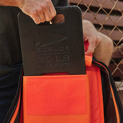 A person is carefully placing a black 20 lb GORUCK ruck plate into an orange GORUCK Rucker 4.0 bag, preparing for another intense rucking session.