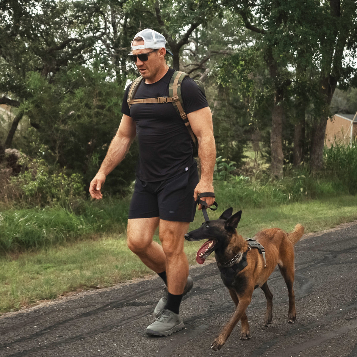 A man wearing a black t-shirt and shorts, paired with a white cap, confidently walks on the road with his brown dog on a leash. He sports sunglasses and carries a backpack while striding in his GORUCK Mackall - Earth + Forged Iron trail shoes, as trees and grass line the roadside behind him.