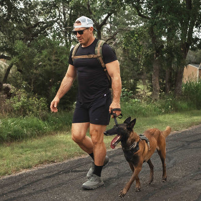 A man wearing a black t-shirt and shorts, paired with a white cap, confidently walks on the road with his brown dog on a leash. He sports sunglasses and carries a backpack while striding in his GORUCK Mackall - Earth + Forged Iron trail shoes, as trees and grass line the roadside behind him.