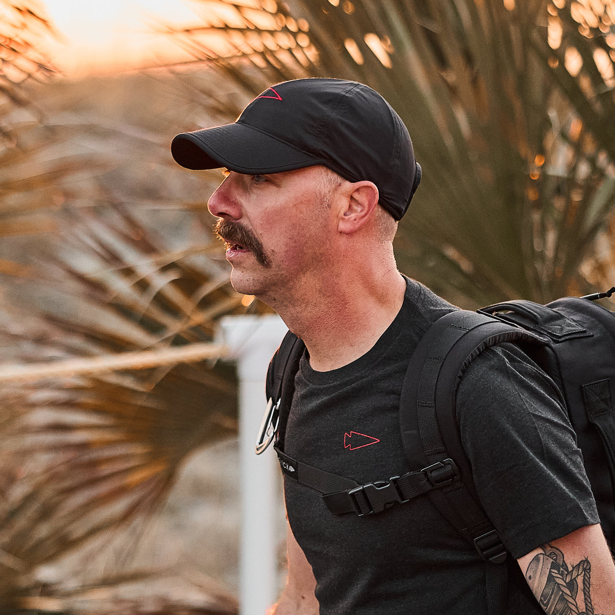 A mustached man wearing a Performance Running Hat - ToughMesh and a backpack walks outdoors with swaying palm leaves in the background.