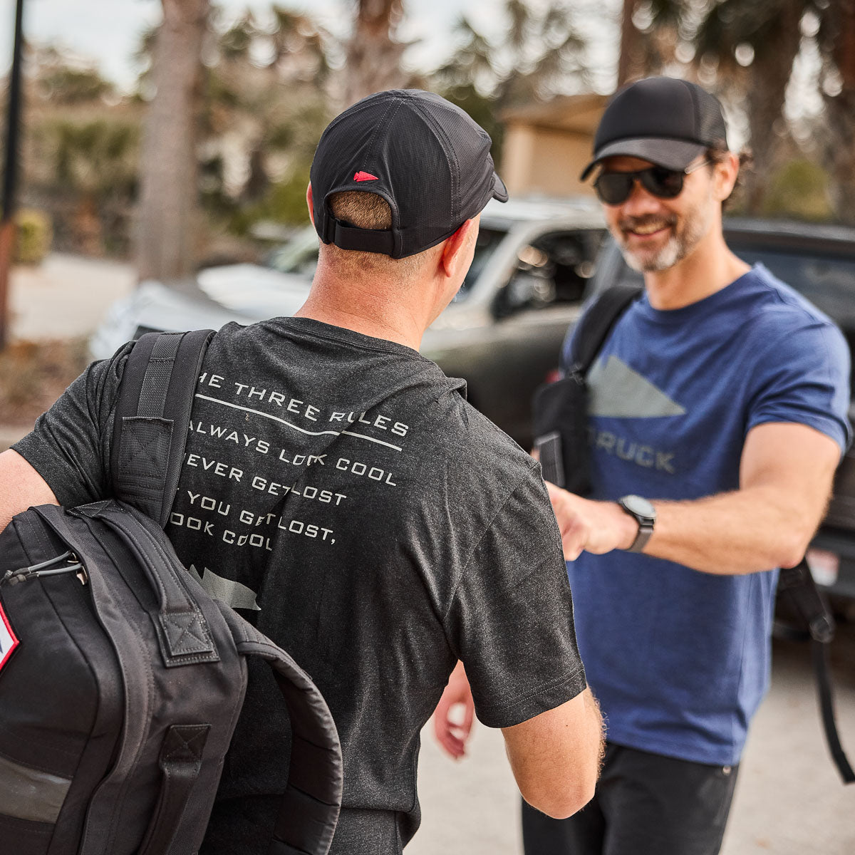 Two men outdoors in casual athletic wear and caps, one showcasing the Performance Running Hat - ToughMesh and carrying a bag, talking and laughing amid a lively backdrop.