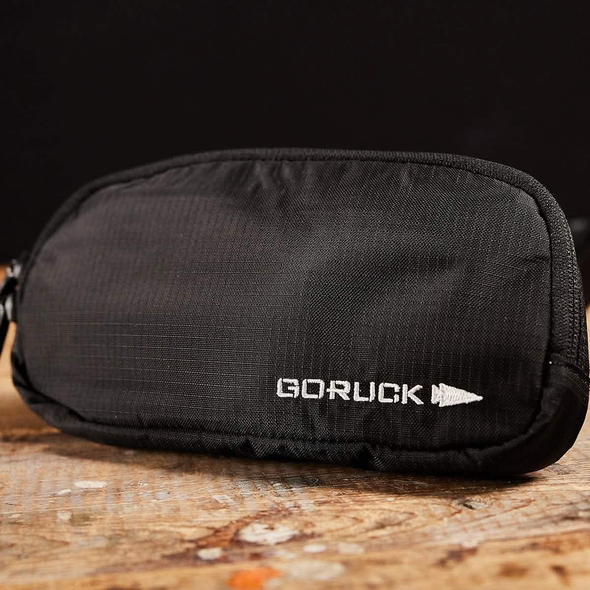 A black Belt Bag - Ripstop ROBIC® by GORUCK, made from durable Robic nylon, is placed on a wooden surface against a dark background. The logo is prominently displayed in white text with a stylized arrowhead design. Its waterproof YKK AquaGuard zippers ensure secure protection from the elements.