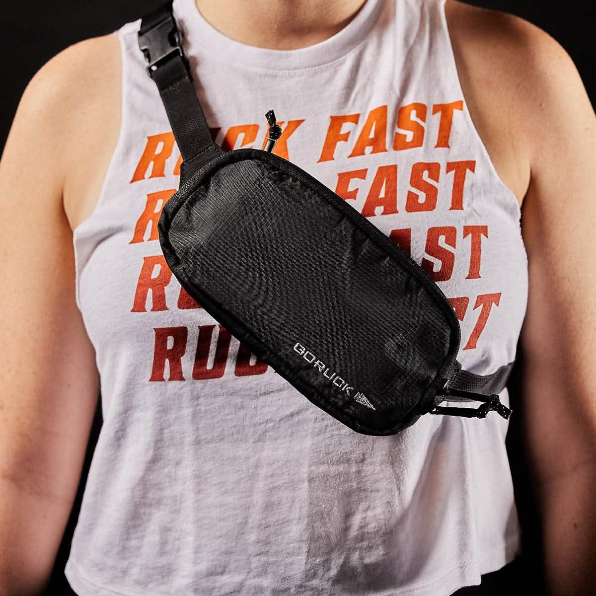 An individual is wearing a sleeveless white shirt with "RUN" repeated in red text, paired with a small black crossbody bag featuring the GORUCK logo. The Belt Bag - Ripstop ROBIC®, positioned at the front across their chest, is equipped with waterproof YKK AquaGuard zippers.