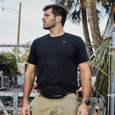 A man in a black t-shirt and khaki pants stands outside, wearing a GORUCK Belt Bag - Ripstop ROBIC®. He has a beard, and the bag is equipped with waterproof YKK AquaGuard zippers. Behind him are palm trees, a chain-link fence, and blurred objects.