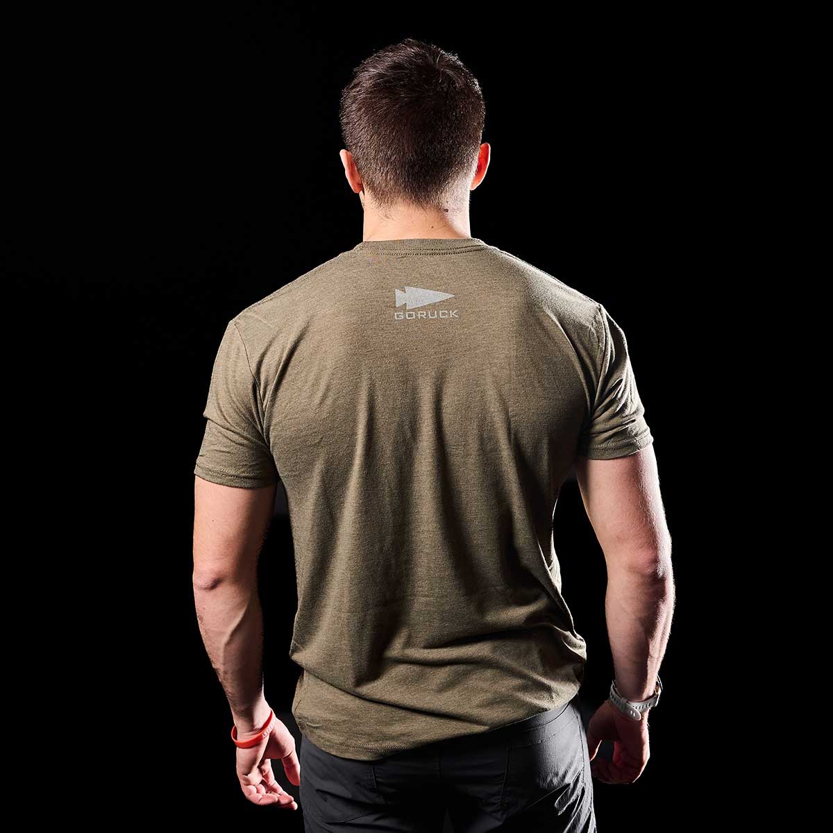 A person dressed in the GORUCK Spearhead Tee - Tri-Blend, featuring a logo on the back, stands facing away against a black background, showcasing Special Forces roots.