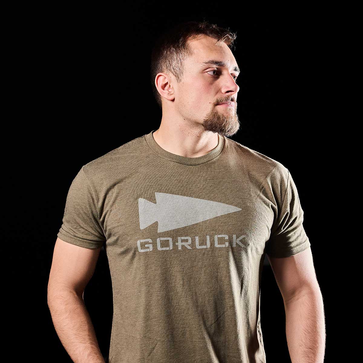 A man sporting a short beard poses against a dark backdrop in an olive green GORUCK Spearhead Tee - Tri-Blend from vendor-unknown. The t-shirt proudly displays the GORUCK spearhead logo, capturing the essence of the brand's Special Forces heritage as he looks to his left.