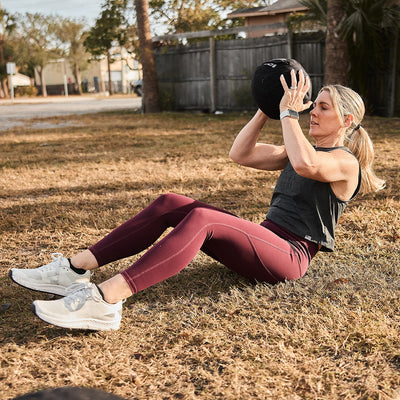 A person is outdoors on a grassy area, performing sit-ups while holding a medicine ball. They are dressed in the Women's Rough Runner outfit by GORUCK, featuring a gray tank top and maroon leggings. For footwear, they sport the Light Grey + Dark Grey high mileage running shoes with determination in their workout. Residential houses and a fence provide the backdrop to their dedicated routine.