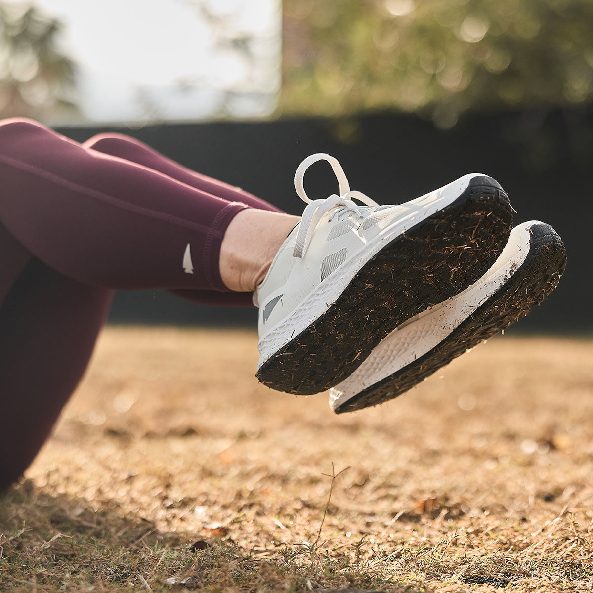 Close-up of a person lying on the grass, wearing GORUCK Women's Rough Runner shoes in light grey and dark grey with burgundy leggings. The focus is on these high-mileage running shoes, equipped with a Gradient Density™ EVA Midsole, set against a blurred background of grass and trees in soft sunlight.