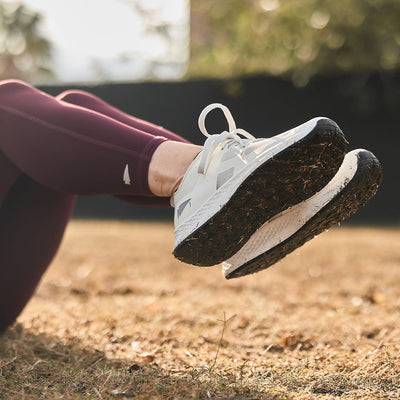 Close-up of a person lying on the grass, wearing GORUCK Women's Rough Runner shoes in light grey and dark grey with burgundy leggings. The focus is on these high-mileage running shoes, equipped with a Gradient Density™ EVA Midsole, set against a blurred background of grass and trees in soft sunlight.