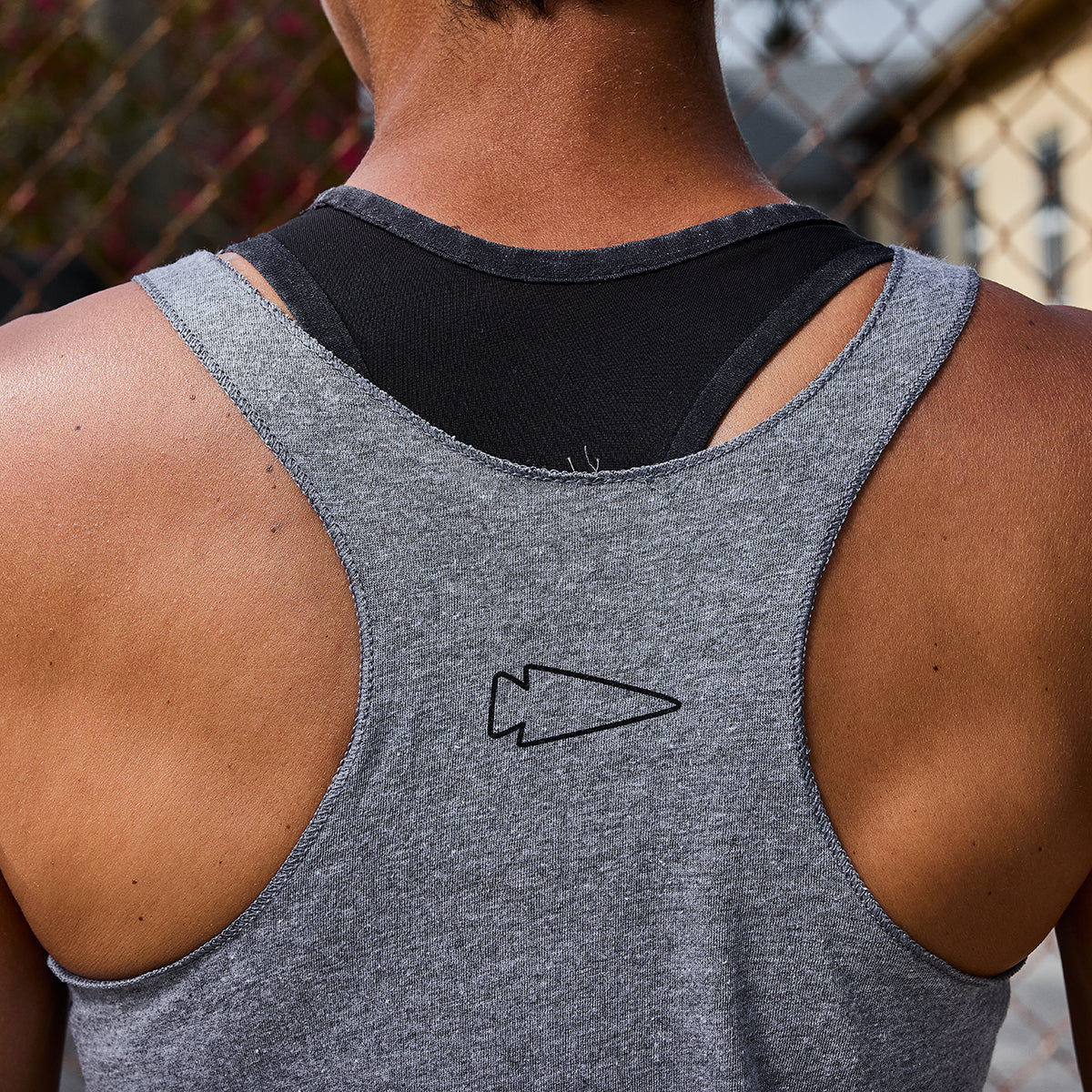 A woman in a Women's Racerback Tank - Tri-Blend and black underlayer stands outdoors by a chain-link fence, ready for an intense workout.