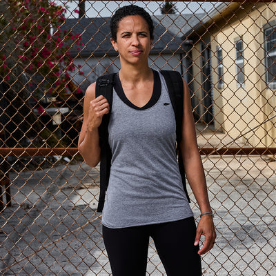 A person stands by a chain-link fence, wearing a Women's Racerback Tank - Tri-Blend in grey and carrying a backpack, with houses visible behind them.