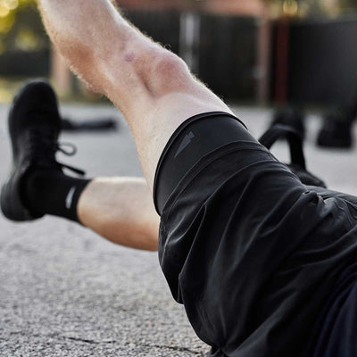 A close-up of someone exercising outdoors shows them wearing GORUCK's Performance Brief and athletic shoes. The focus is on their leg, raised mid-workout, with a blurred pavement and dark objects in the background. The breathable Performance Brief ensures comfort as they move through their routine.