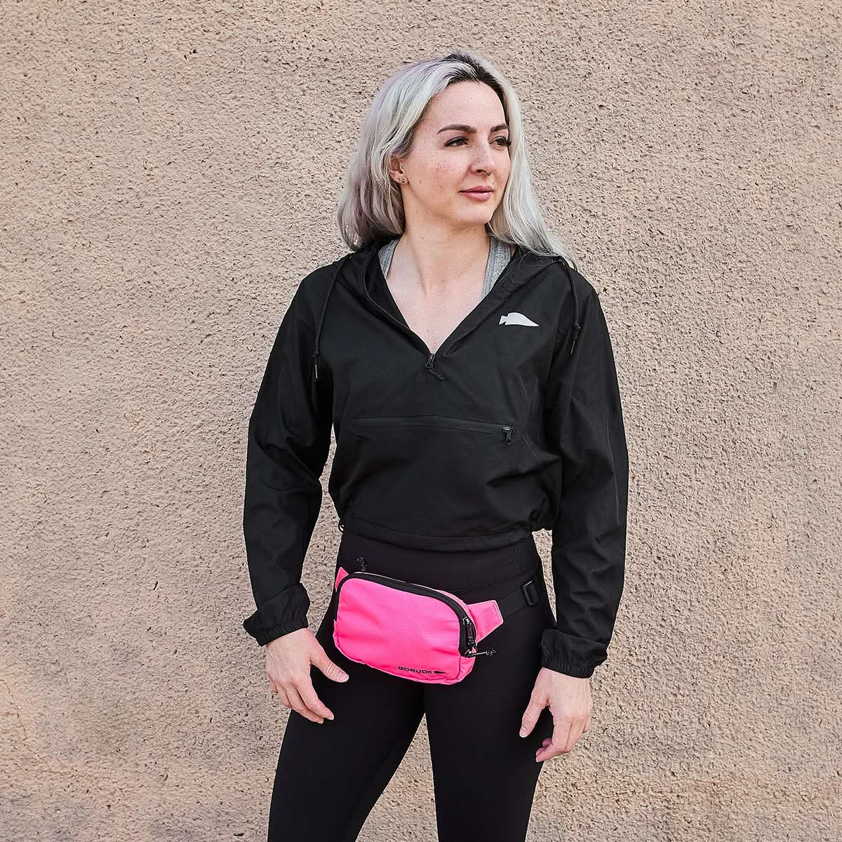 A person with long hair stands against a textured wall, wearing a black jacket and leggings. They sport a chic pink Belt Bag - Ripstop ROBIC® by GORUCK around their waist, featuring Waterproof YKK AquaGuard zippers for added flair and functionality.