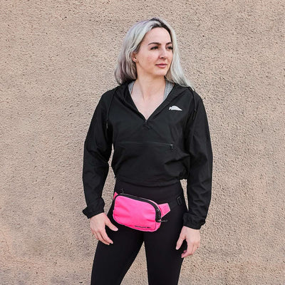 A person with long hair stands against a textured wall, wearing a black jacket and leggings. They sport a chic pink Belt Bag - Ripstop ROBIC® by GORUCK around their waist, featuring Waterproof YKK AquaGuard zippers for added flair and functionality.