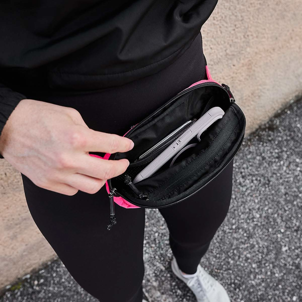 A person dressed in black is holding a pink GORUCK Belt Bag - Ripstop ROBIC®, which contains a white smartphone and features Waterproof YKK AquaGuard zippers, while standing on textured pavement.