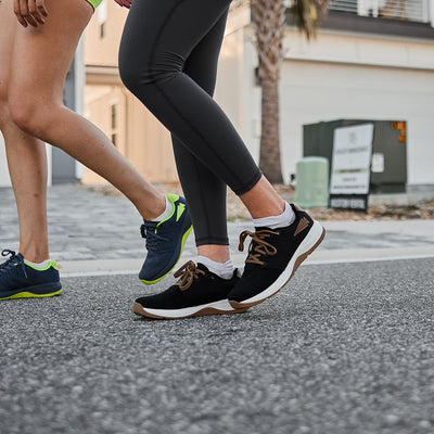 Two individuals are walking on a street, their legs visible. One of them is wearing gray leggings paired with GORUCK's Women's Ballistic Trainers in Black + White with Coyote Reflective Spearhead accents. The other person is in shorts and vibrant blue and neon green sneakers. A building and a sign can be seen in the background.