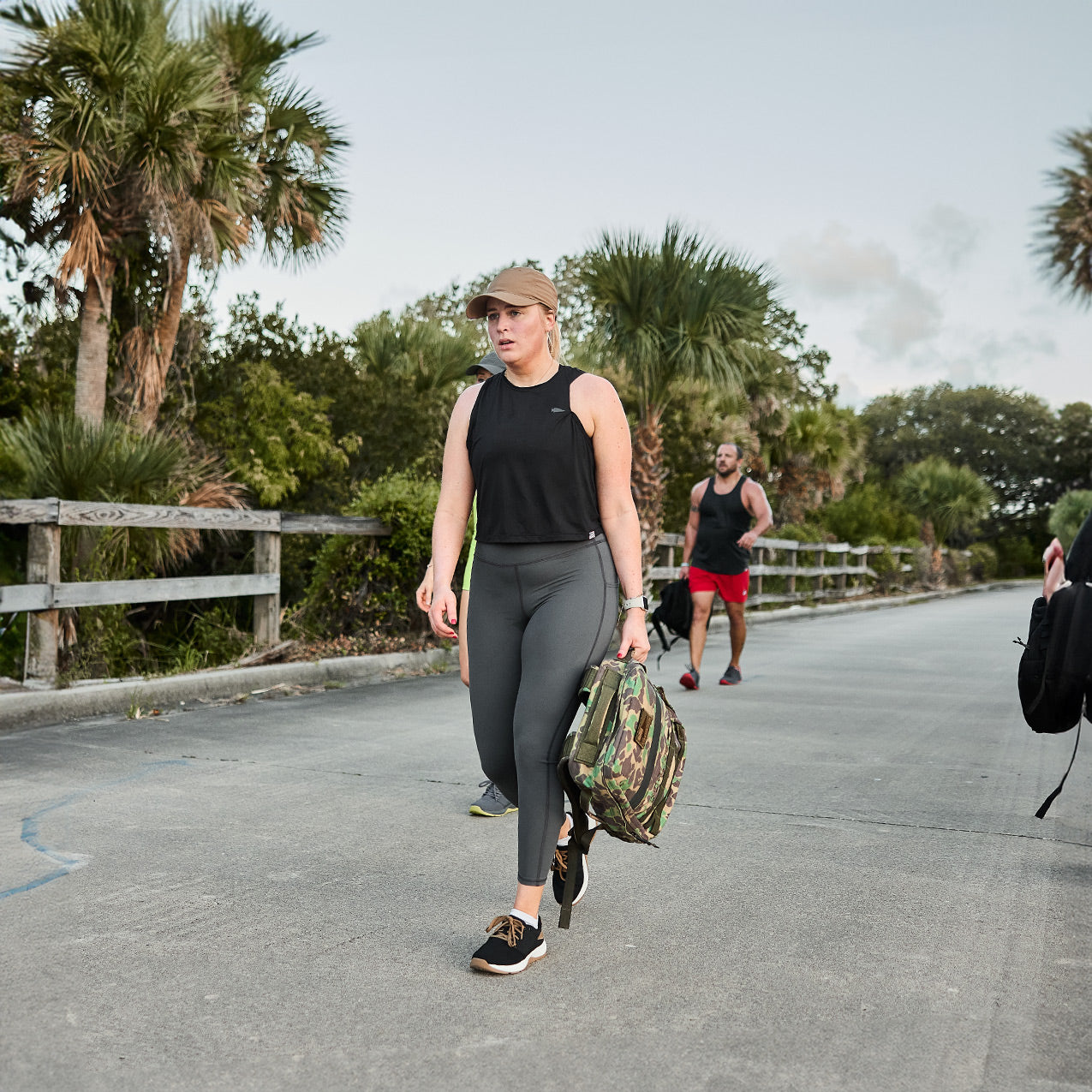 A woman dressed in athletic clothing sports a GORUCK Women's Ballistic Trainers - Black + White with Coyote Reflective Spearhead, carrying a CORDURA Ballistic Nylon backpack as she strolls down a palm tree-lined road. In the distance, a man jogs in a tank top and red shorts under an overcast sky.