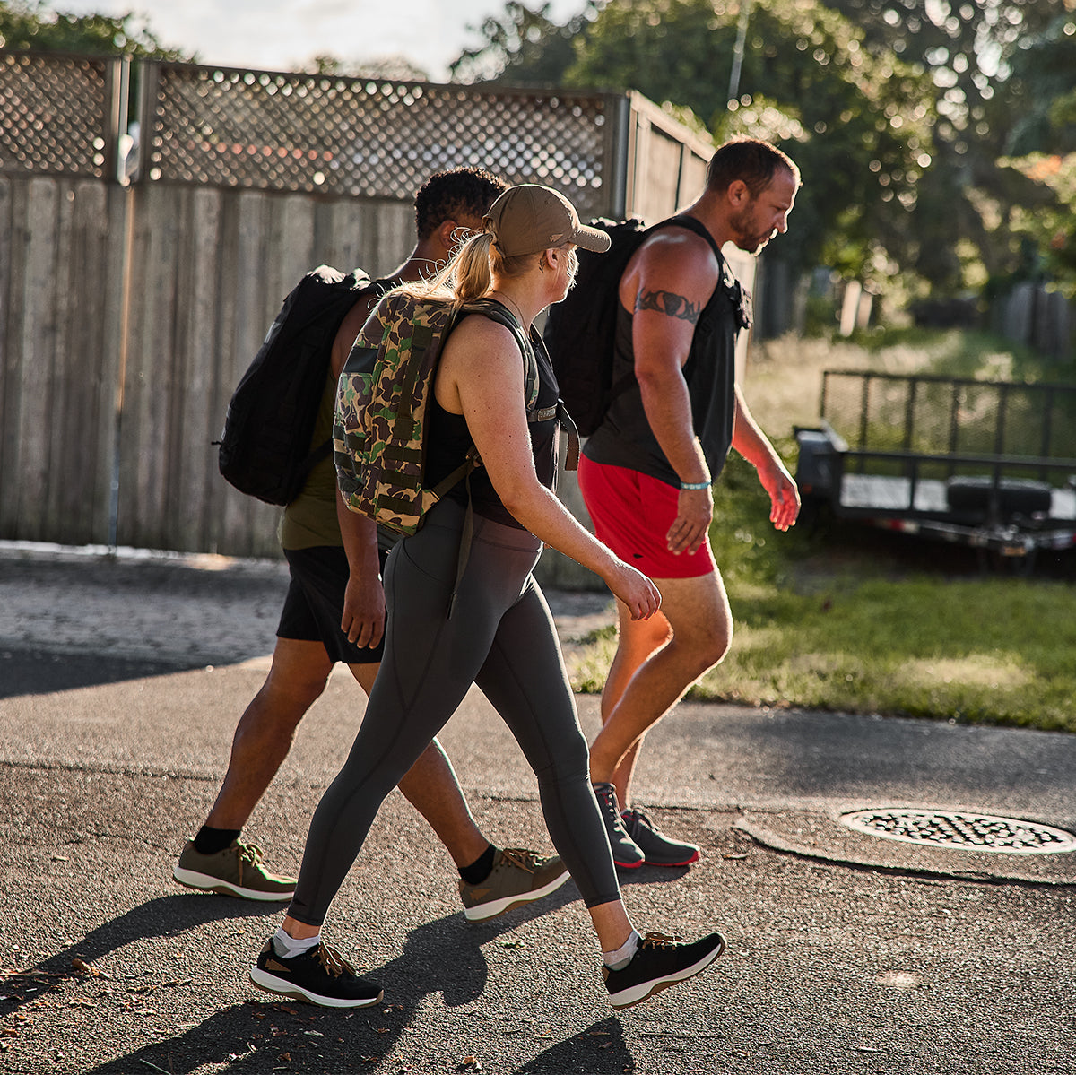 Three people are walking outdoors on a paved path, dressed in athletic clothing and carrying backpacks. Wearing the Women's Ballistic Trainers in Black + White with Coyote Reflective Spearhead by GORUCK, they stride confidently under the sunny sky, surrounded by a wooden fence and lush greenery.