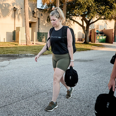 A woman in athletic gear strolls down the street carrying a weighted bag, sporting a black tank top, green shorts, and GORUCK's Women's Ballistic Trainers in Ranger Green + White with Coyote Reflective Spearhead. Her backpack, made from durable CORDURA® Ballistic Nylon, complements her active lifestyle. Trees and a building stand in the background.